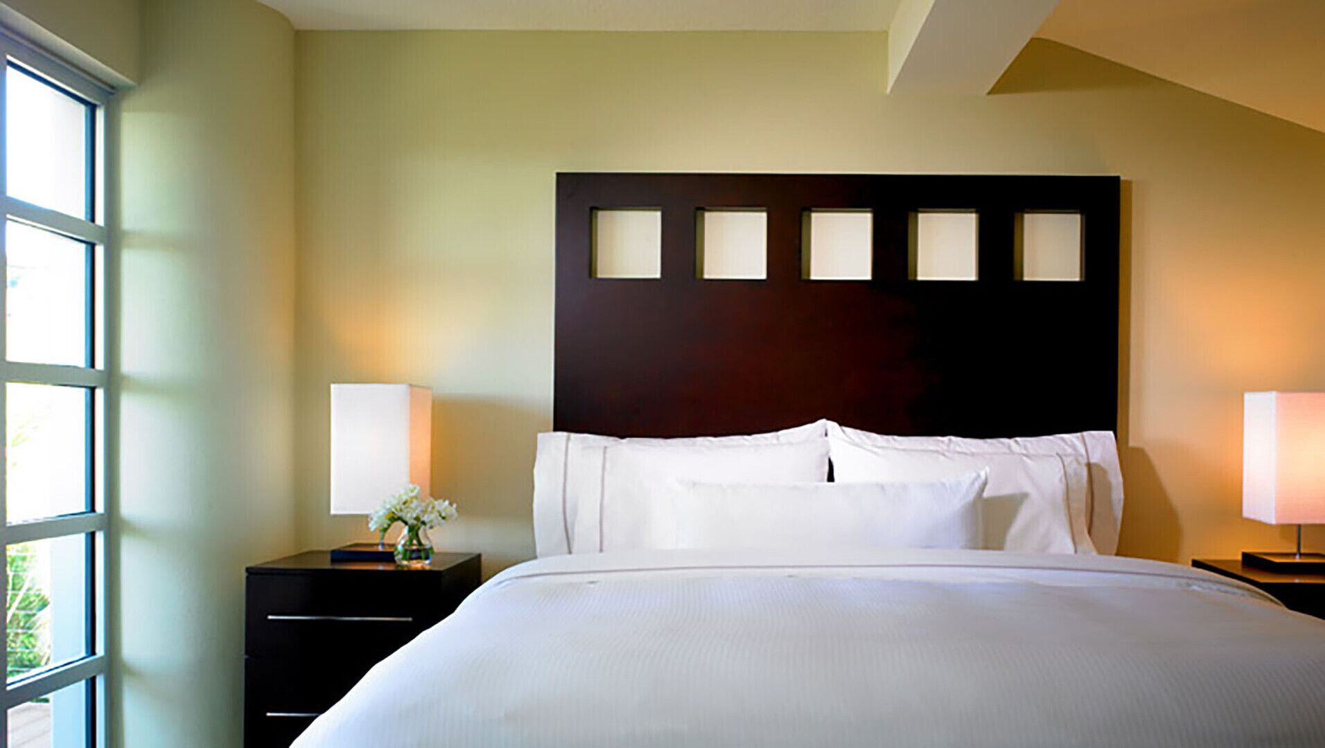 A hotel room with a large bed, two bedside tables, a dark headboard, and a window on the left.