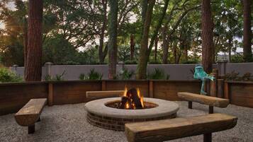 A fire pit with benches, surrounded by a gravel area and wooden walls, with trees in the background.
