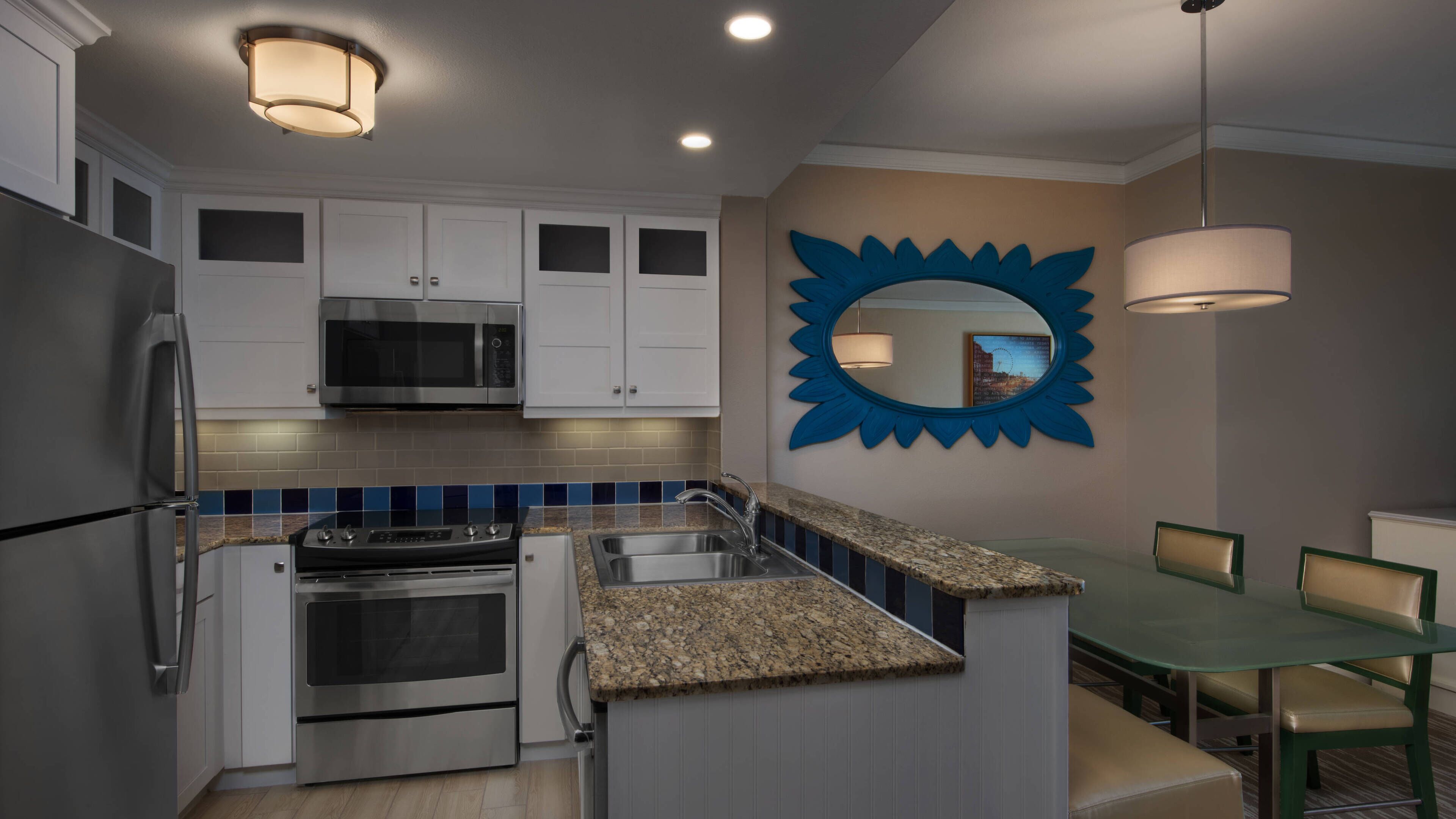 Una cocina moderna con armarios blancos, electrodomésticos de acero inoxidable, una encimera de granito y un comedor con mesa y sillas de vidrio.
