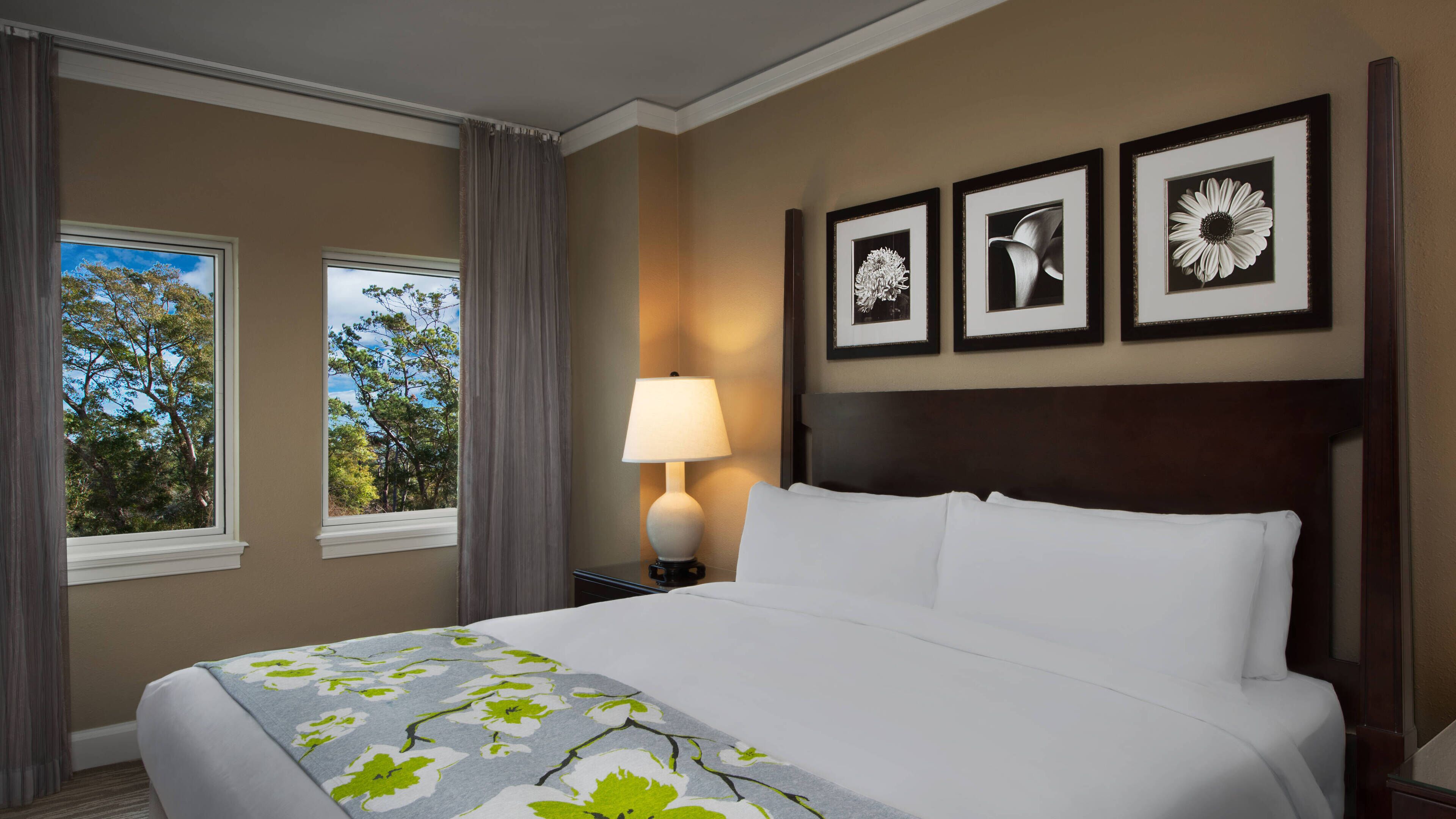 Ein Hotelzimmer mit einem großen Bett, einem Nachttisch mit Lampe, drei gerahmten Bildern an der Wand und einem Fenster mit Vorhängen.