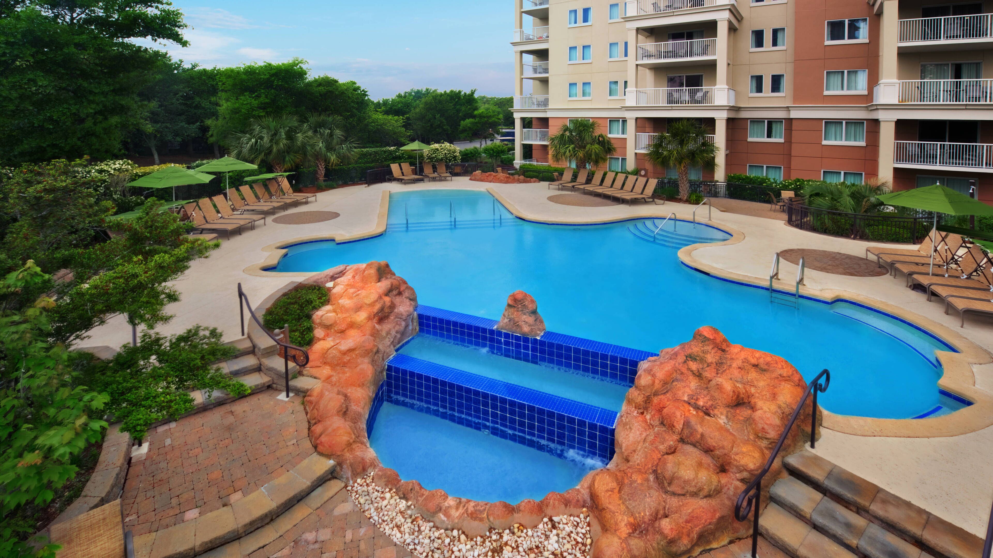 A swimming pool with a rock feature, surrounded by lounge chairs and umbrellas, with a multi-story building in the background.