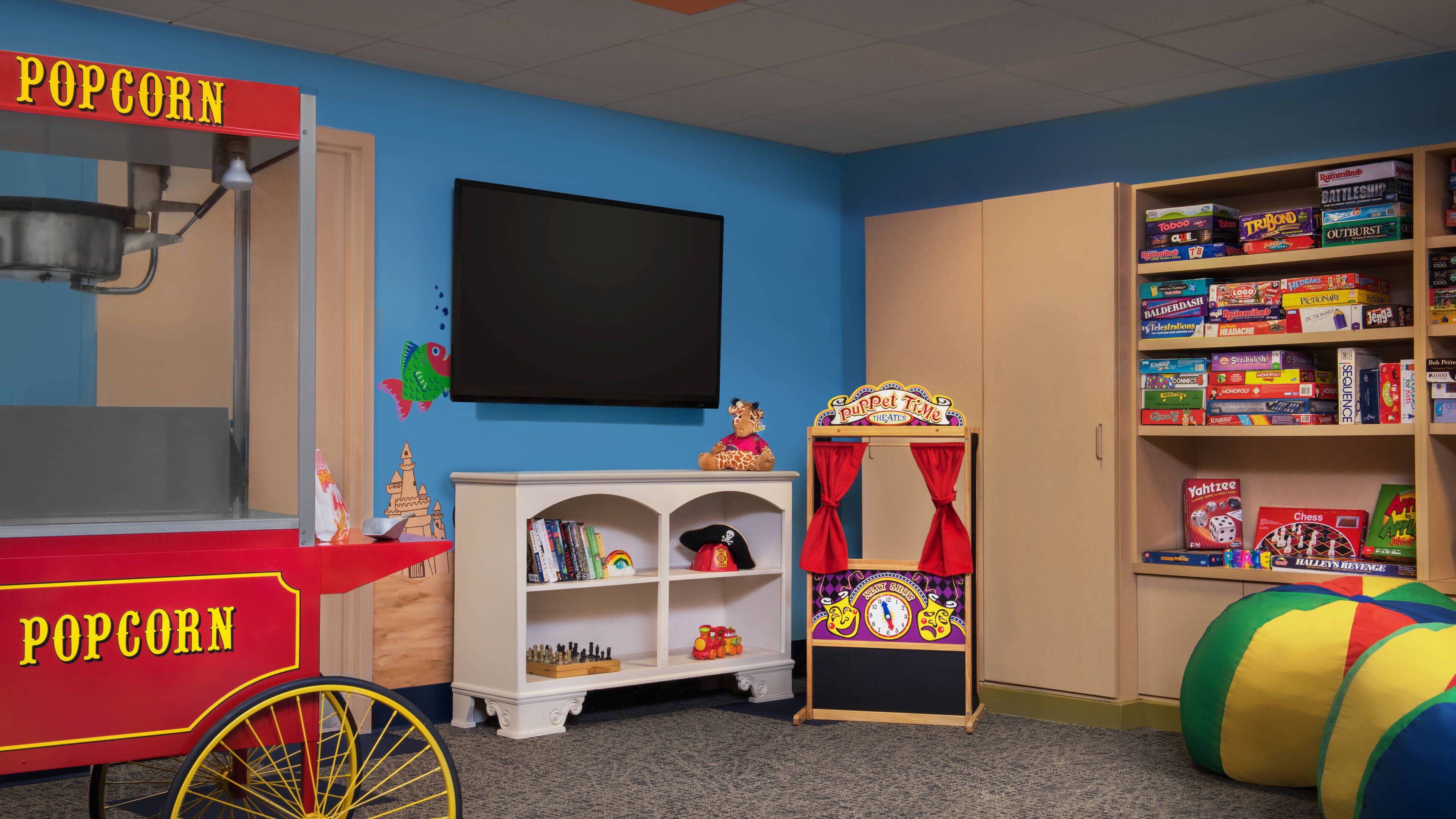 Une salle de jeux pour enfants avec un chariot à pop-corn, une bibliothèque remplie de jeux de société et un théâtre de marionnettes.