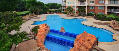 A swimming pool with a rock feature, surrounded by lounge chairs and umbrellas, with a multi-story building in the background.