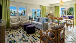 A hotel room with a sofa, armchairs, a dining table, and chairs. There are windows with a view of palm trees and the ocean.