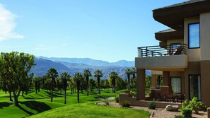 Un moderno edificio de dos pisos con balcón, rodeado de un césped cuidado y palmeras, con vistas a un paisaje montañoso.