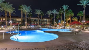 Un área de piscina nocturna con sillones y sombrillas, rodeada de palmeras y un edificio con balcones.