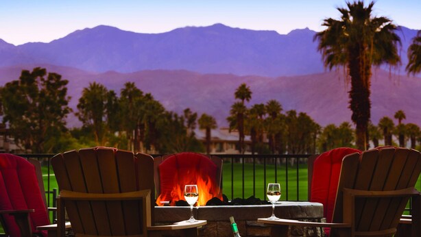 Una terraza con sillas y mesas de madera, una chimenea y vistas a montañas y palmeras.