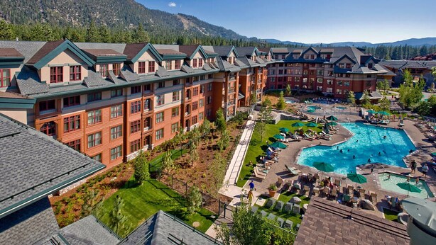 Un resort con varias edificaciones, un área de piscina central con tumbonas y un paisaje montañoso de fondo.