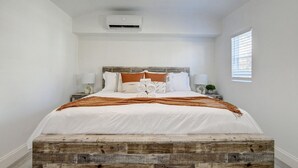 A bedroom with a wooden platform bed, white bedding, and a small bedside table with a lamp. - Bliss Near Beach w/ Guest House + Salt Heated Pool (Boynton Beach)