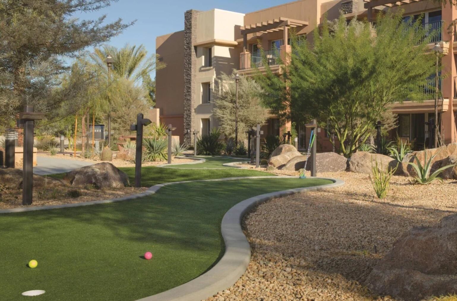 A golf course with a well-maintained green, a putting area, and a pathway leading to a building with multiple balconies.