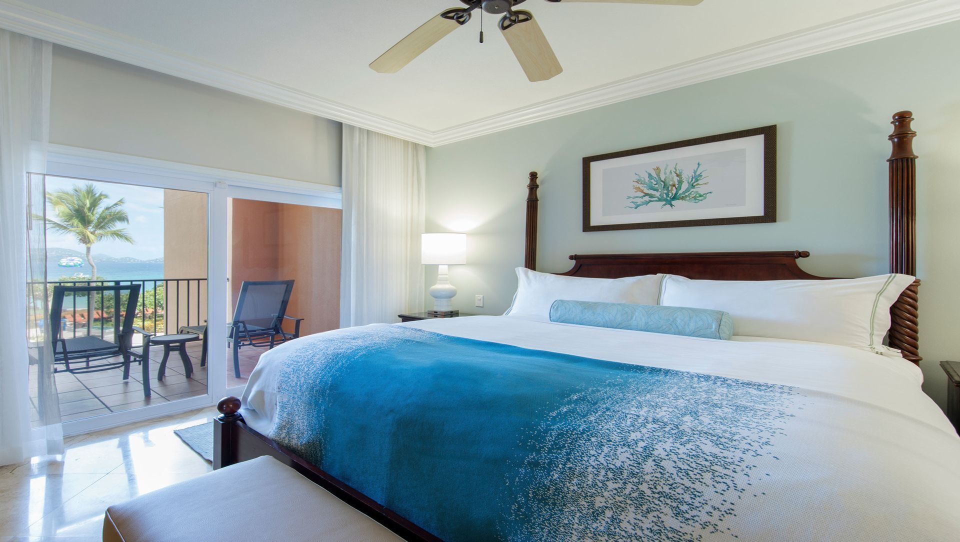 Une chambre d’hôtel avec un grand lit, un ventilateur de plafond, un balcon avec des chaises et une œuvre d’art encadrée au mur.