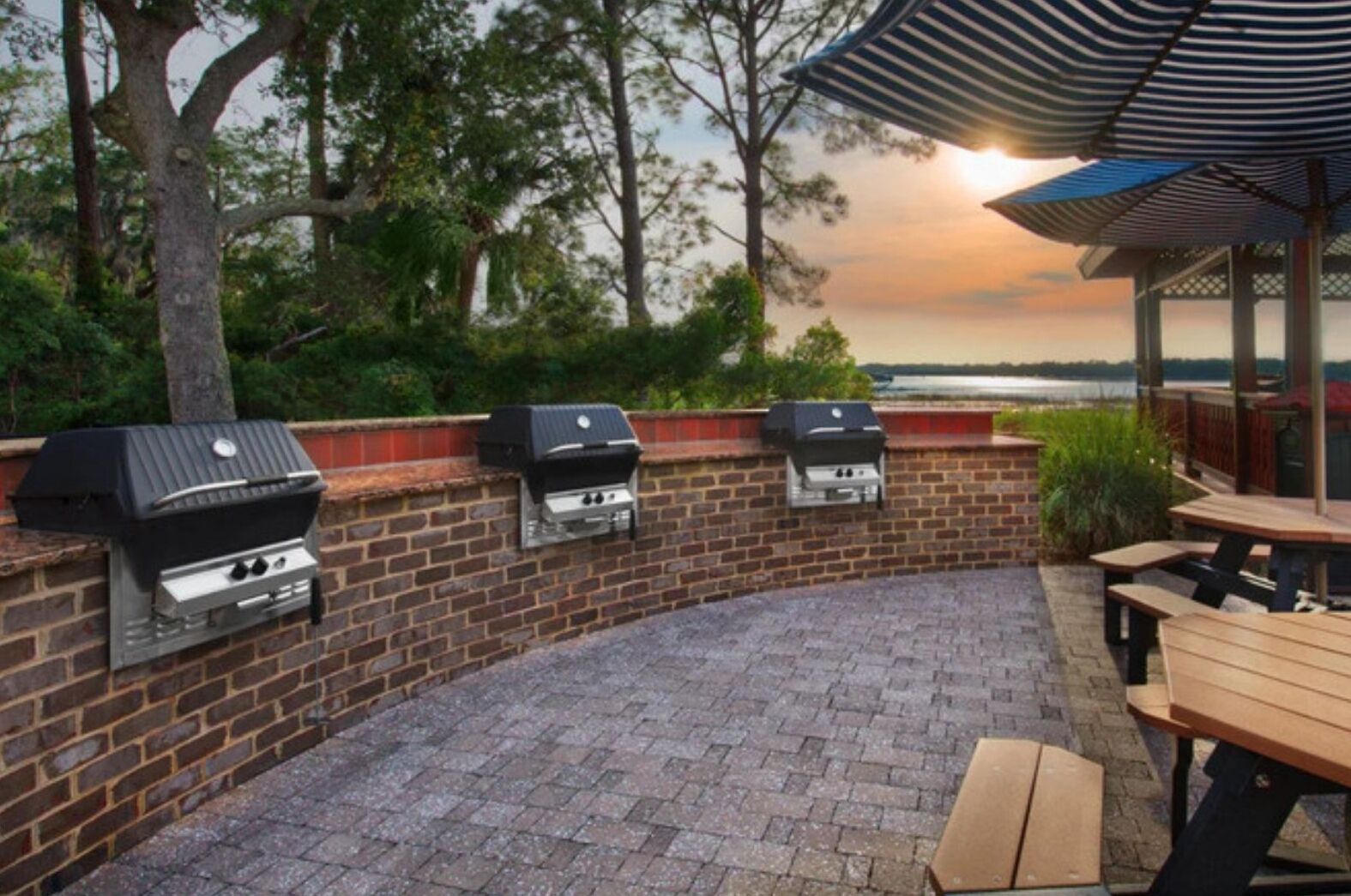 An outdoor patio with brick flooring, three built-in barbecue grills, and wooden picnic tables.