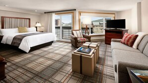 Ein Hotelzimmer mit einem großen Bett, einer Couch, einem Sessel, einem Fernseher und Blick auf Gebäude durch die Fenster.