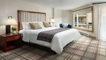 Habitación de hotel con una cama grande, dos lámparas de noche, una mesita de noche con una planta, una ventana con vista a un edificio y una alfombra estampada.