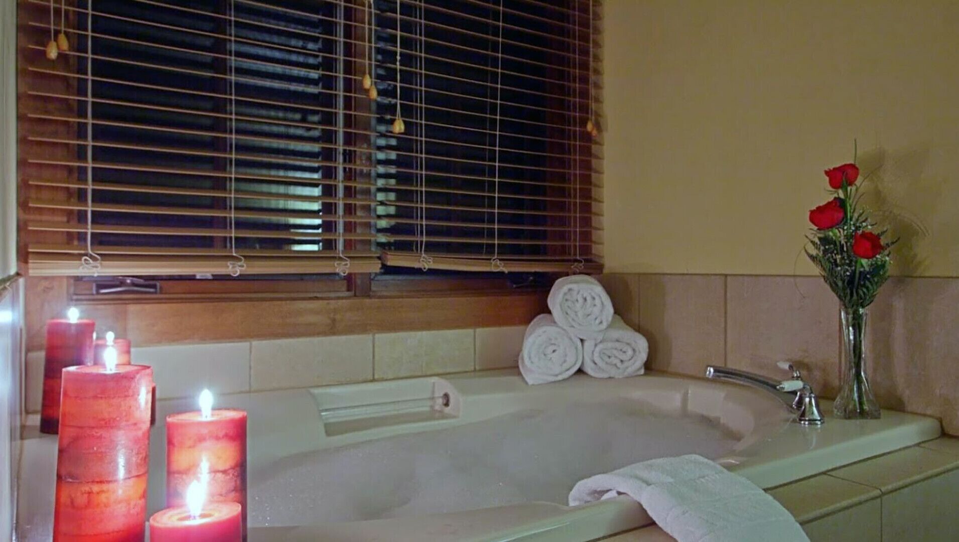 A bathroom with a bathtub, a window with blinds, a vase with red flowers, and two lit candles.