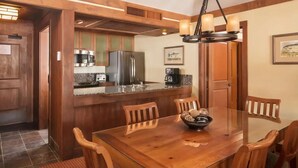 A wooden dining table with chairs, a bowl of snacks, a kitchen area with a refrigerator, stove, and coffee maker.