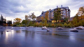 Een modern hotel met meerdere verdiepingen, een rivier op de voorgrond en herfstkleurige bomen.