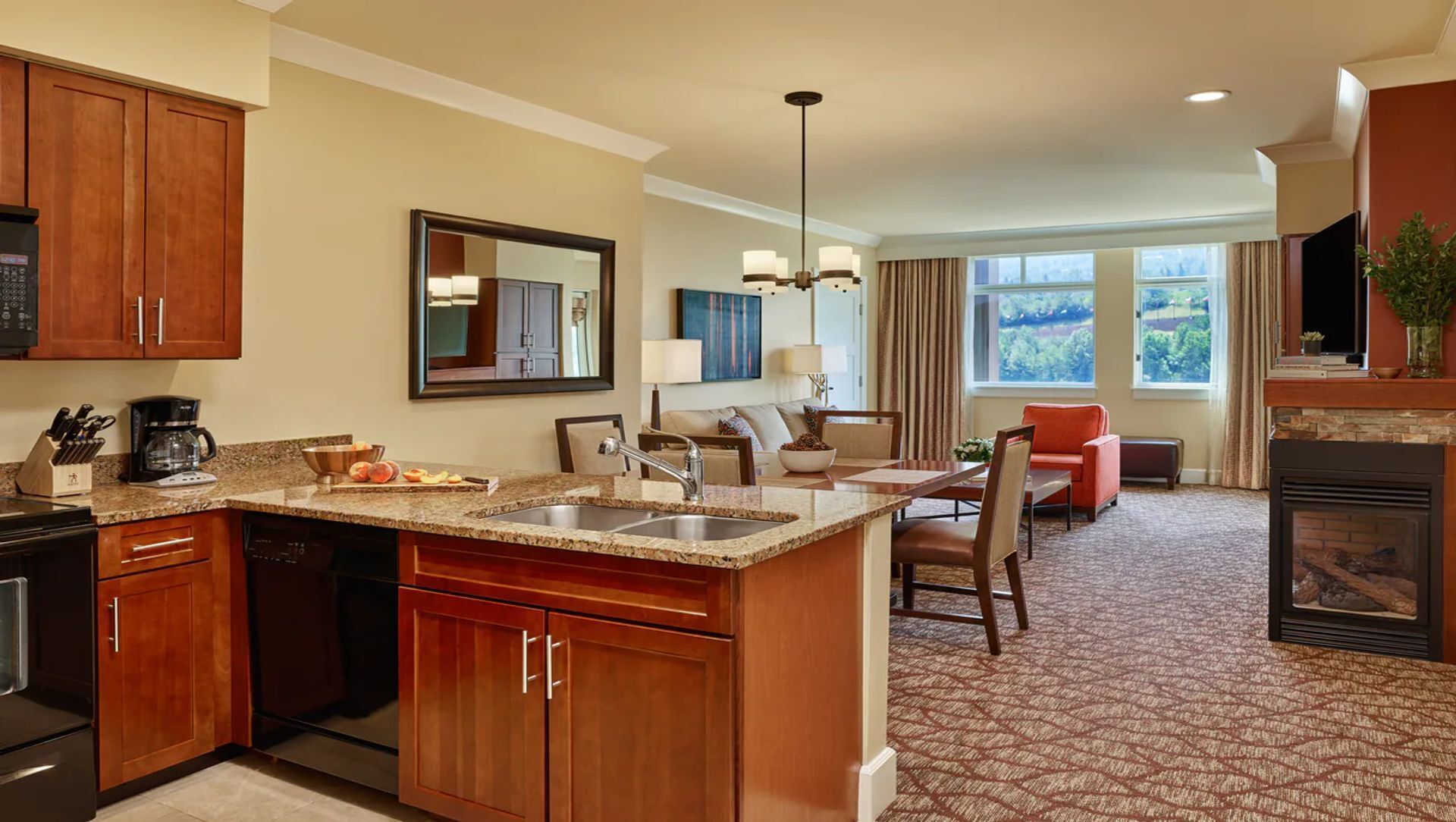A hotel kitchen with granite countertops, a sink, and wooden cabinets. There is a dining area with a table and chairs, and a fireplace.