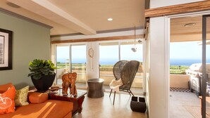 A spacious living area with a large window offering a view of the ocean, a wooden sculpture, a wicker chair, and a grill outside. - Oceanfront Coral Reef (Solana Beach)