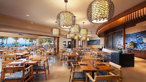 A spacious restaurant with wooden tables and chairs, large hanging lamps, and a view of the ocean.