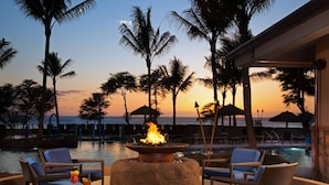 A poolside lounge with wicker chairs, a fire pit, and a view of the ocean at sunset.
