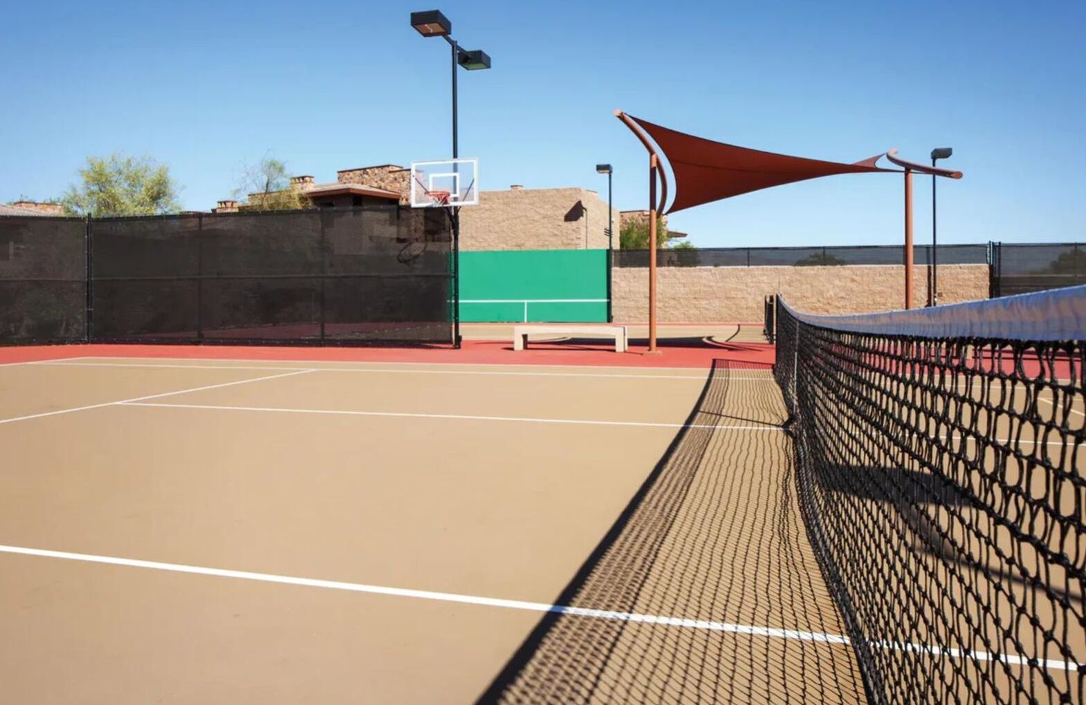 An outdoor tennis court with a net, a basketball hoop, and a shaded area.