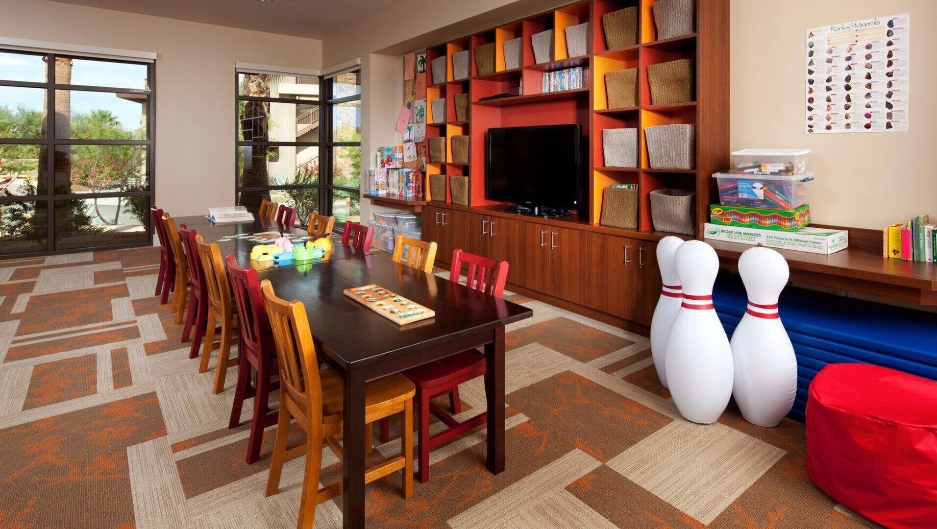 A dining area with a large table, chairs, and a television. There are shelves with baskets and books.