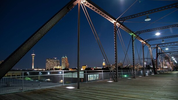 Ein Nachtblick auf eine Brücke mit Holzbelag und Metallgestell, mit Blick auf einen beleuchteten Stadt-Horizont.