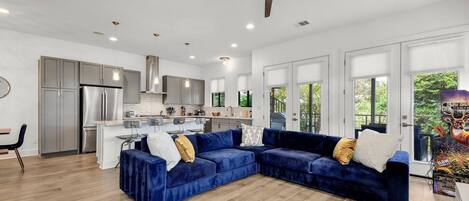 Una sala de estar moderna con un sofá modular azul, una cocina con electrodomésticos de acero inoxidable y un ventilador de techo.