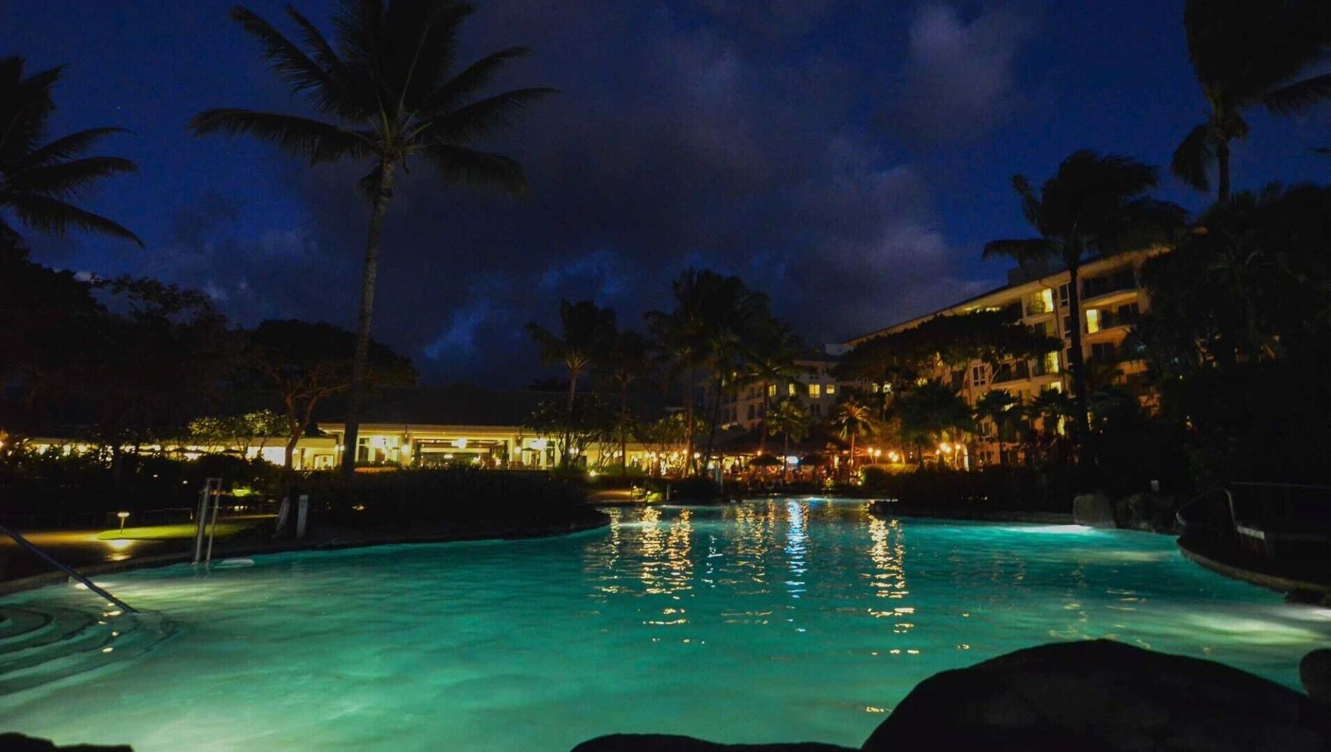 A nighttime view of a hotel pool area with a clear pool, palm trees, and illuminated buildings.