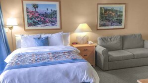 Habitación de hotel con cama, sofá gris, dos cuadros en la pared, mesita de noche con lámpara y mesilla con teléfono.