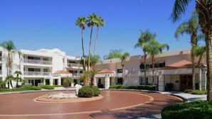 Un hotel de varios pisos con un patio central, palmeras y un cielo azul despejado.