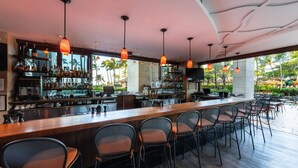 A bar area with multiple seating arrangements, a counter with various bottles and glasses, and a view of palm trees outside.