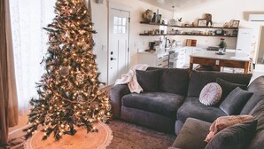 A cozy living room with a decorated Christmas tree, a gray sofa, and a kitchen in the background. - Cottage with gated driveway near downtown & river (Grants Pass)