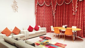 Ein Wohnzimmer mit einem beigefarbenen Sofa, orangefarbenen Kissen, einem runden Couchtisch, einem farbenfrohen Teppich, einem Esstisch mit orangefarbenen Stühlen und roten Vorhängen mit floralem Muster.