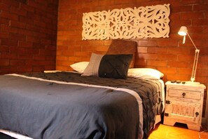 Une chambre avec un mur en briques, une tête de lit décorative, un lit recouvert d’une couverture sombre et une table de chevet avec une lampe.