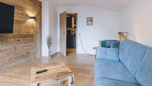 Een woonkamer met een houten wand, een blauwe bank, een salontafel en een aan de wand bevestigde televisie.