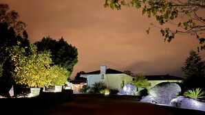 A well-lit residential property at night with a driveway leading to a house, surrounded by landscaped gardens and trees.