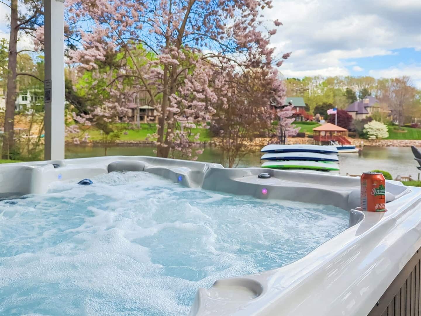 A hot tub with a can of Sunkist juice on the edge, surrounded by a deck with a view of a lake and cherry blossoms.