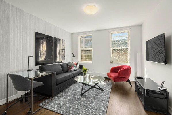 A modern living room with a black sofa, a red chair, a glass coffee table, and a TV on the wall.
