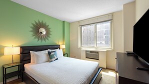 Ein ordentlich bezogenes Bett mit weißer Bettwäsche, eine grüne Wand, ein dekorativer Spiegel, ein Fenster mit Jalousien und ein Flachbildfernseher auf einem Schreibtisch.