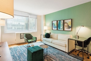 A modern living room with a green patterned wall, a white sofa, a green armchair, a small round table, and a glass desk with a chair.
