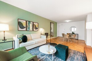 A modern living room with a white sofa, green armchair, and a round coffee table. There is a dining area with a table and chairs, and a kitchen area with a stove and cabinets.