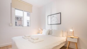 A neatly made bed with white linens and towels, a bedside table with a lamp, a framed artwork on the wall, and a window with a view of buildings.