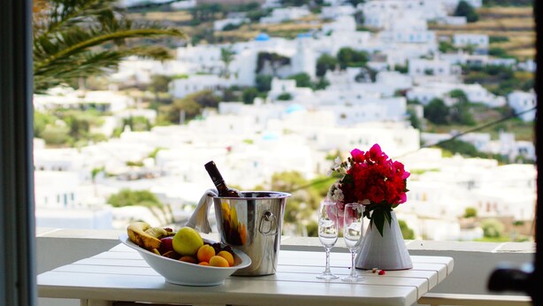 Uma mesa com uma fruteira, um recipiente de metal e um vaso com flores, com vista para uma cidade caiada de branco.