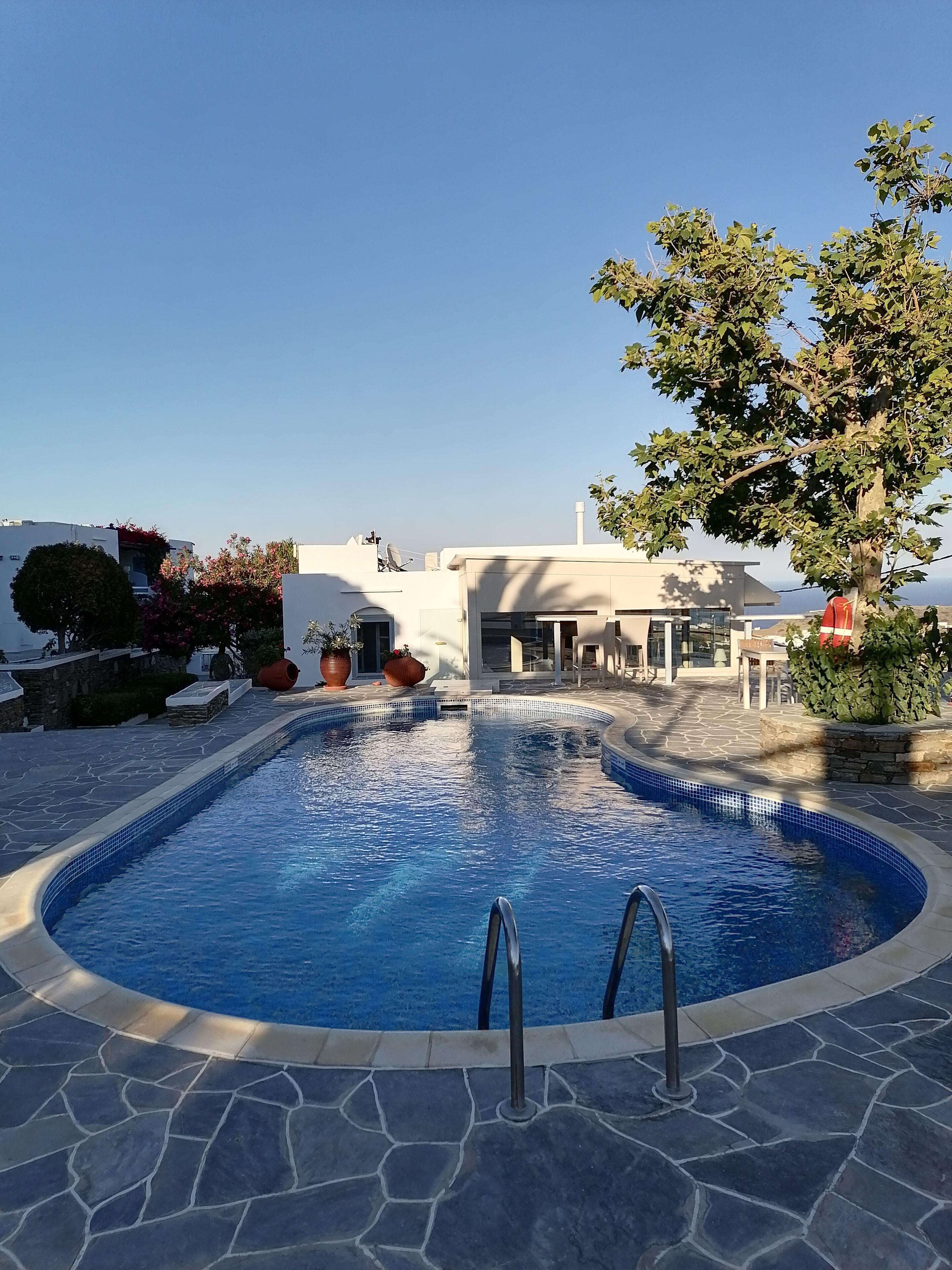 Une piscine aux bords recouverts de pierres, entourée d’une terrasse en pierre, et un bâtiment à la façade blanche en arrière-plan.
