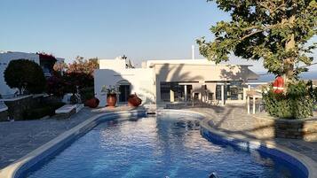 Une piscine aux bords recouverts de pierres, entourée d’une terrasse en pierre, et un bâtiment à la façade blanche en arrière-plan.