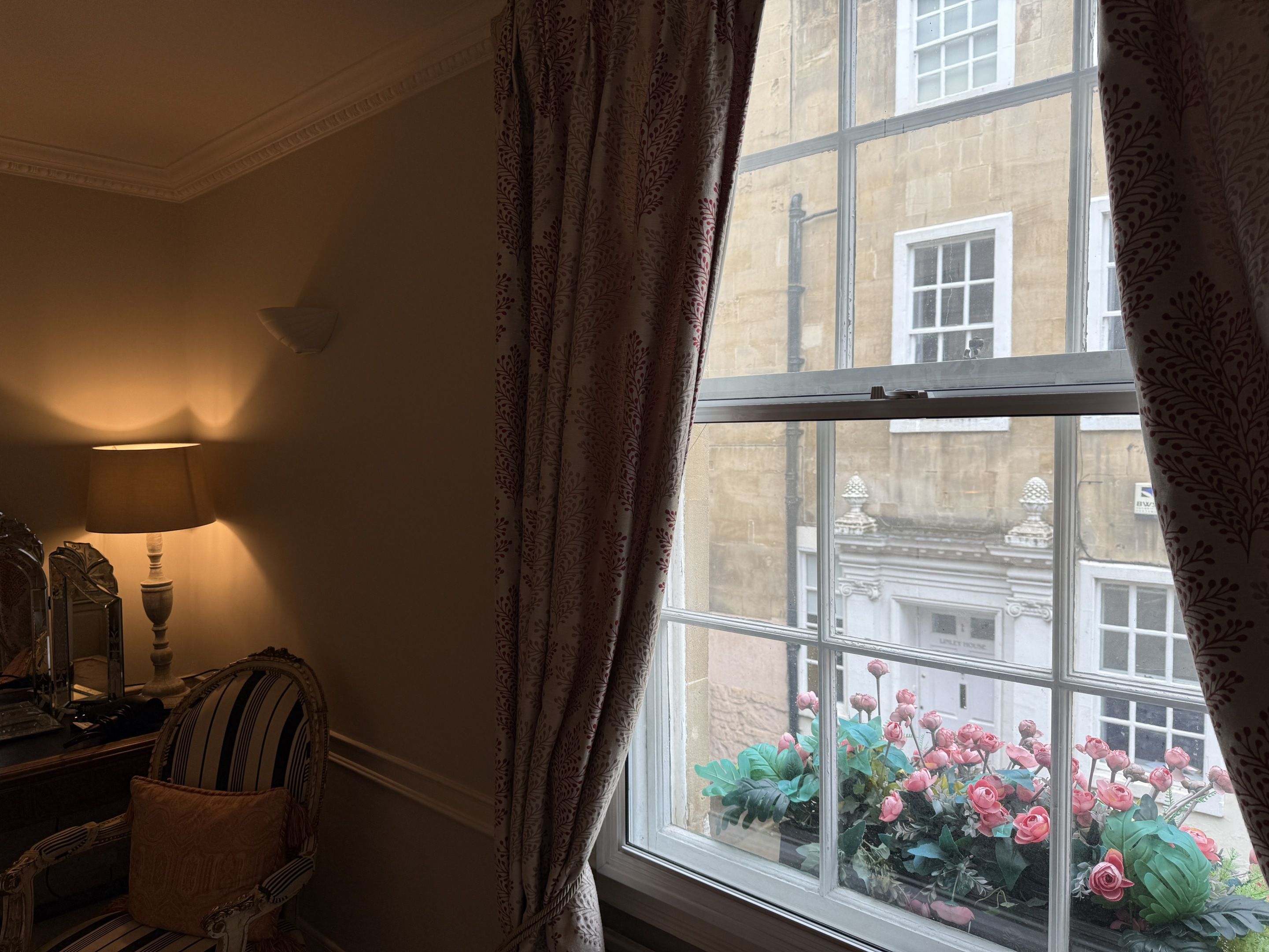 Una habitación con ventana, lámpara, sillón y vista a un edificio con macetas florales.