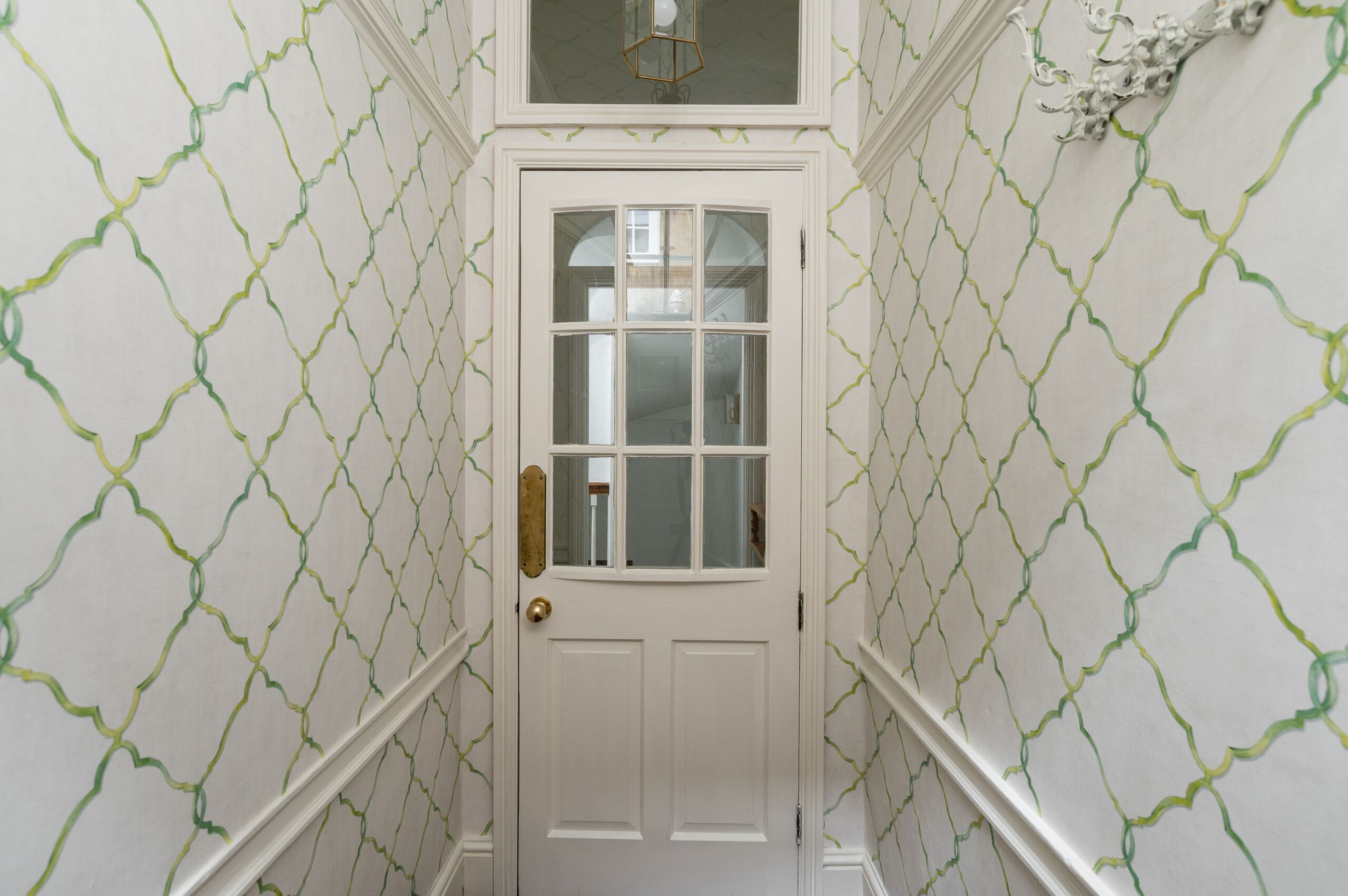Un pasillo estrecho con una puerta blanca, un papel tapiz con diseño verde y blanco, y un adorno en la pared sobre la puerta.