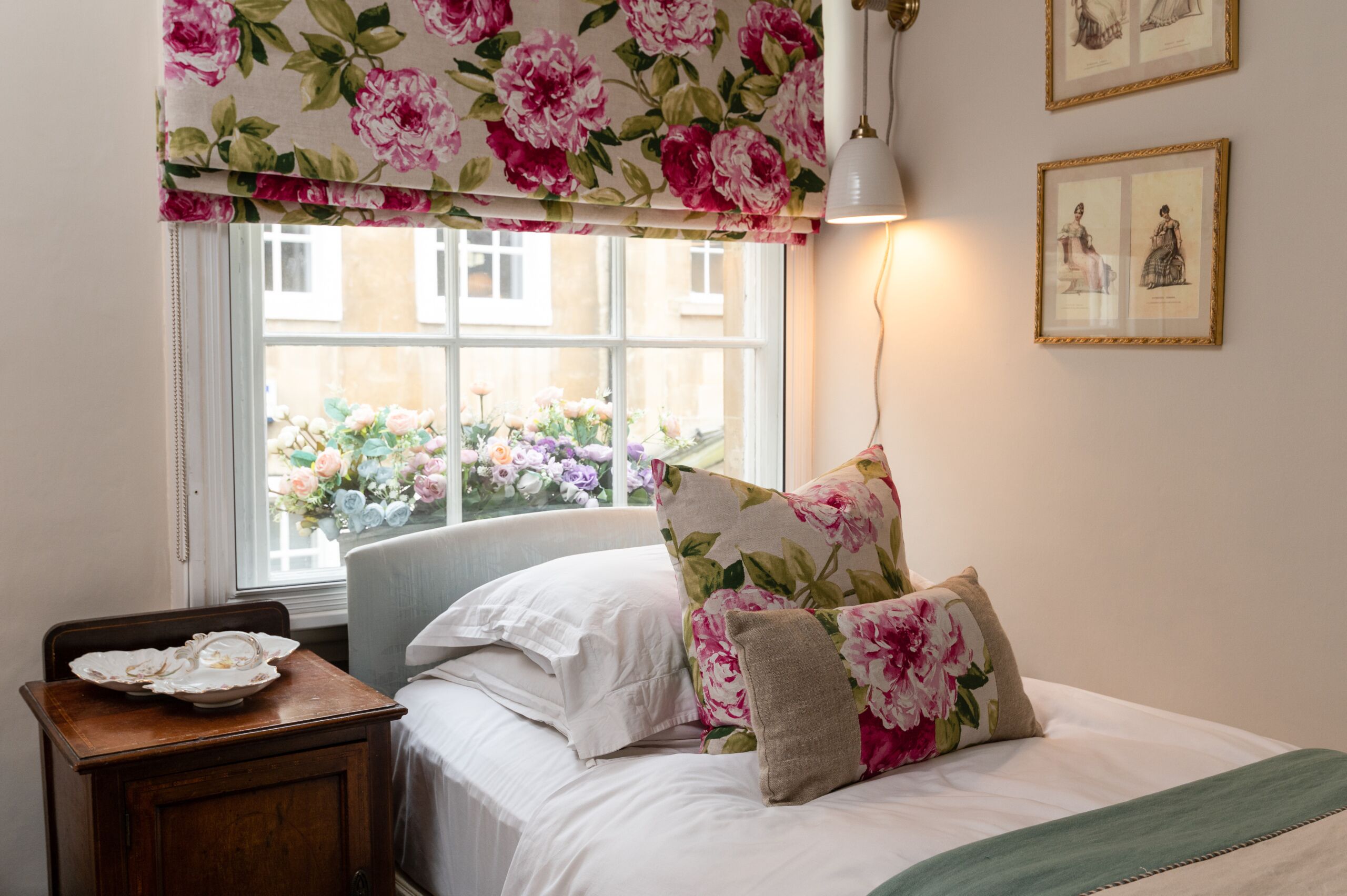 Un dormitorio con una cama, una mesita de noche, una ventana con cortinas florales y cuadros en la pared.